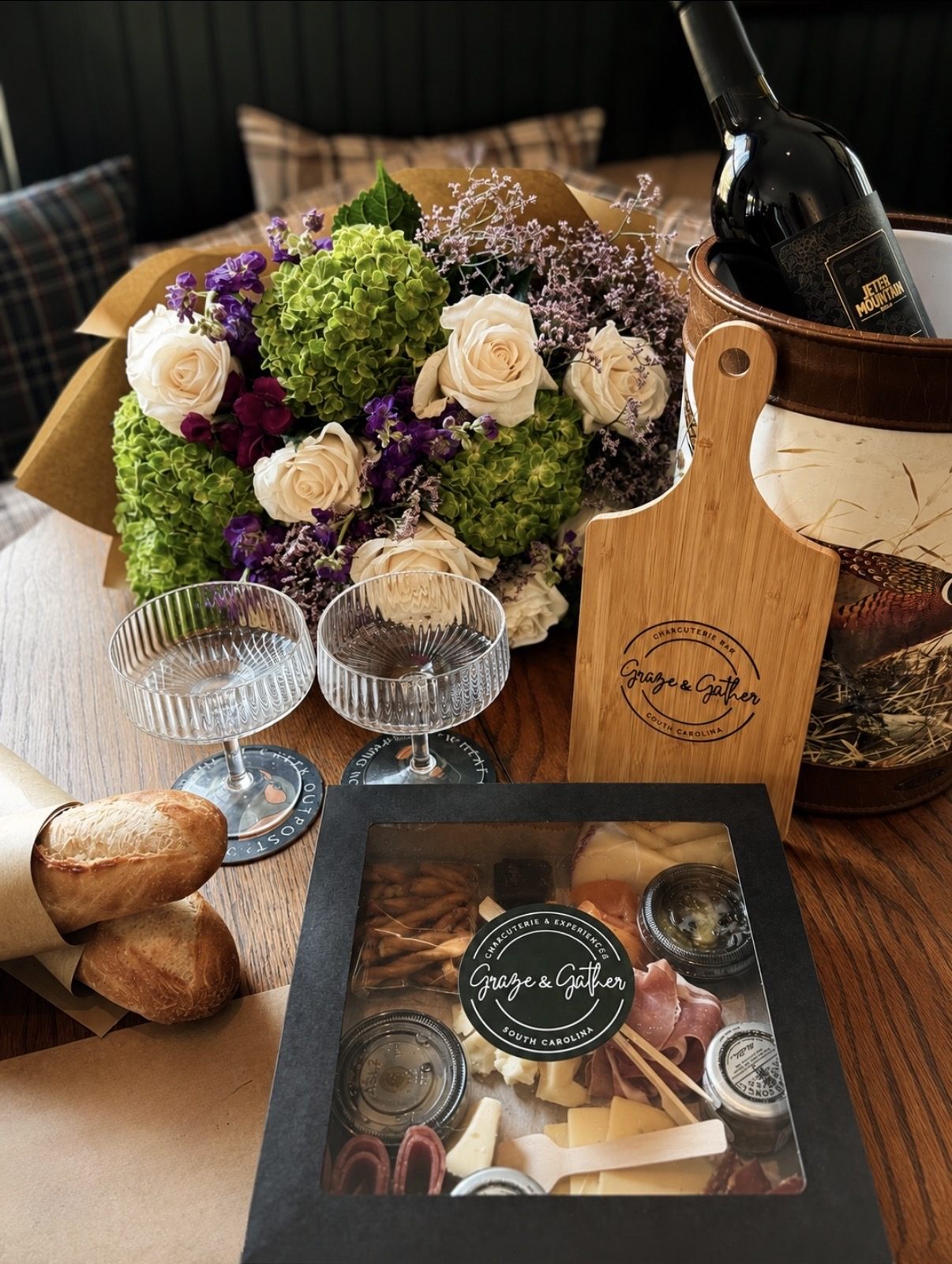 A window-lid Graze & Gather box filled with cheeses, prosciutto, breadsticks and preserves, set beside a branded serving board, wine and fresh flowers
