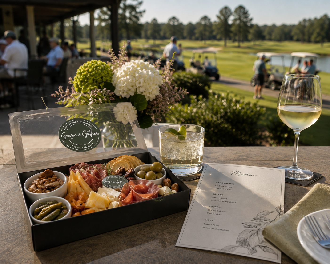 A Graze & Gather charcuterie box open on a clubhouse terrace beside a glass of wine, with the course and carts behind