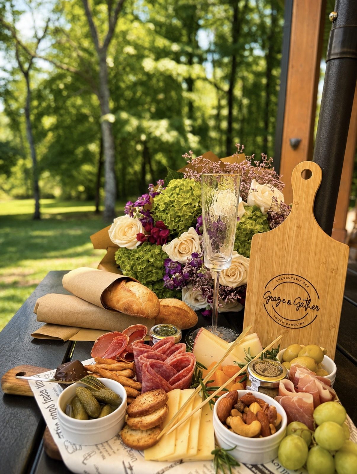 A Graze & Gather board laid out on an outdoor table with a branded bamboo serving board, champagne and fresh flowers